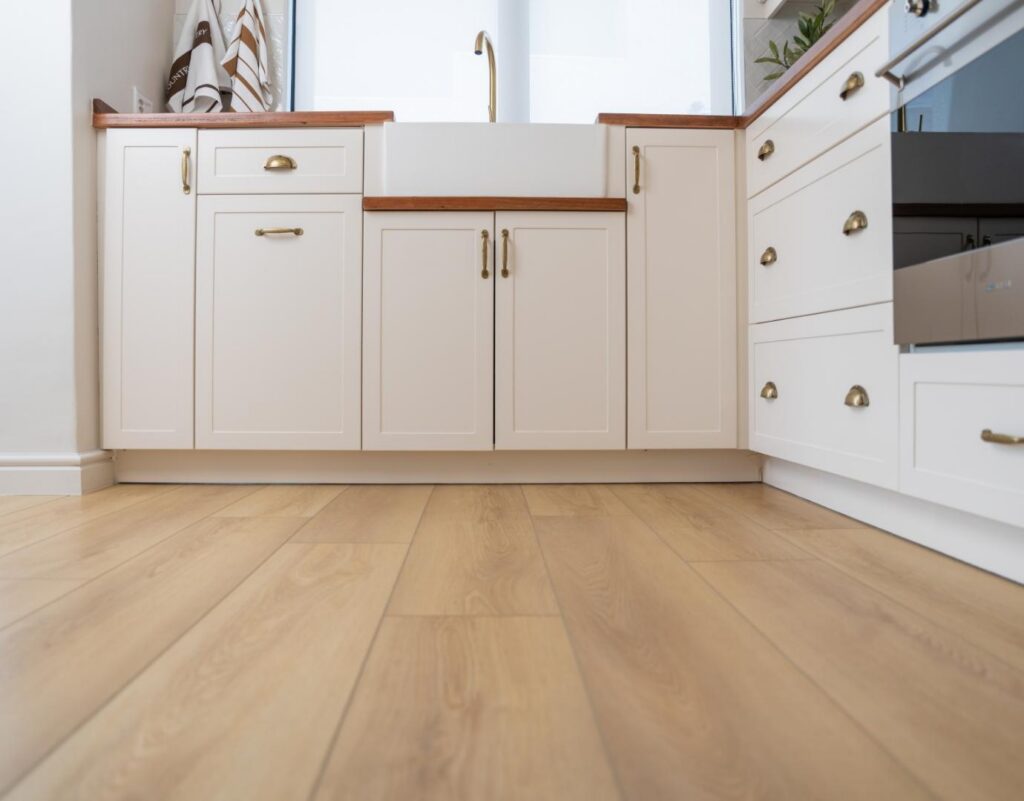 Low-angle close-up view of light natural oak luxury vinyl plank flooring installed in a kitchen with white shaker-style cabinets, brass handles, and a farmhouse sink.