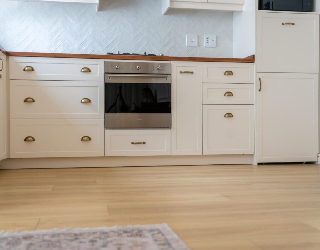 Eye-level view of light oak vinyl flooring running along a modern kitchen with white shaker drawers, a built-in stainless steel oven, white herringbone tile backsplash, and a patterned area rug in the foreground.