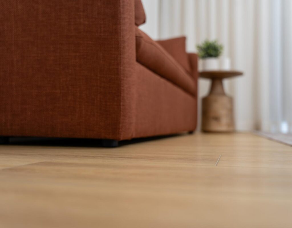 Close-up detail shot of light-colored luxury vinyl plank flooring next to the base of a rust-orange upholstered couch and a small wood side table.
