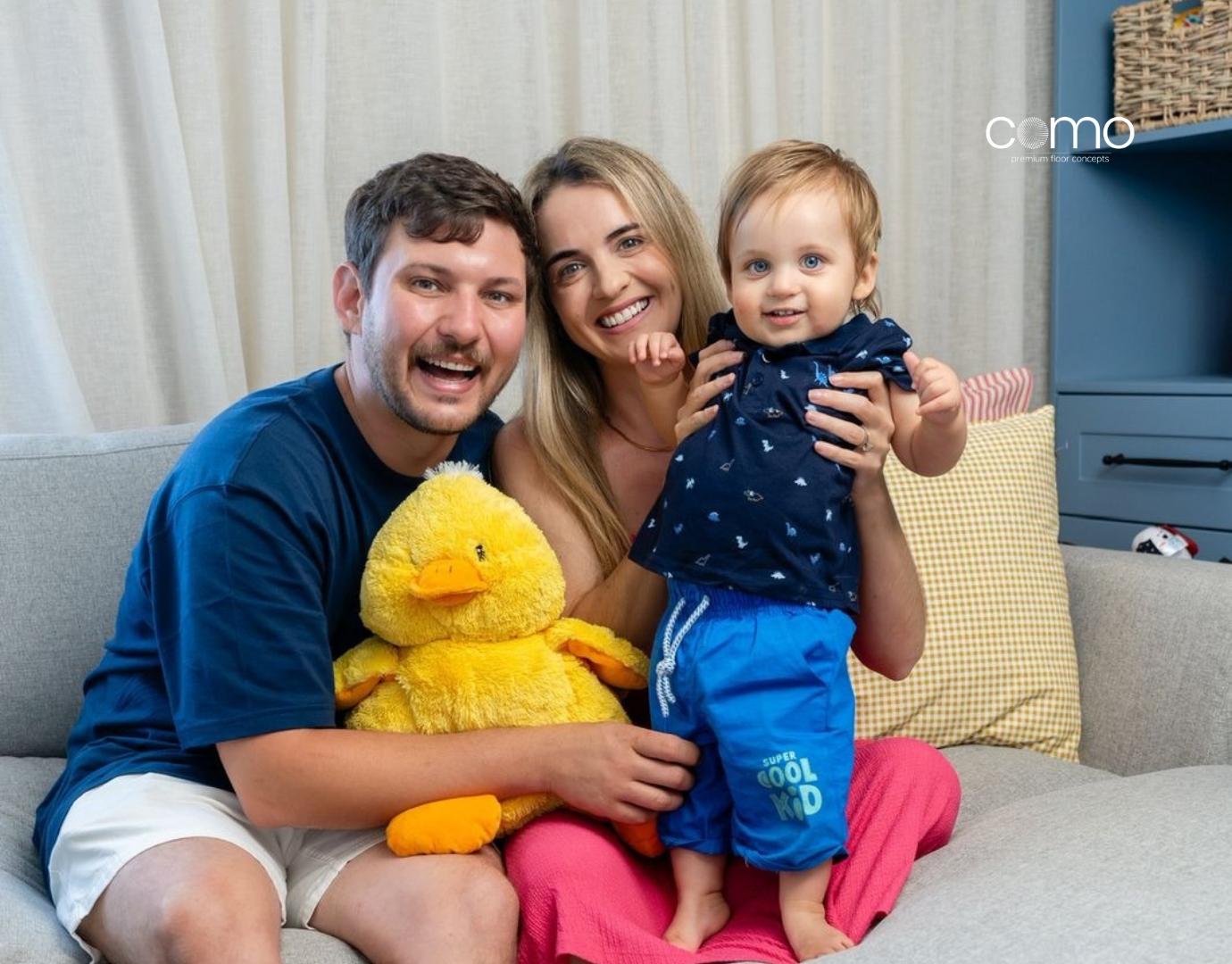 Blog Post Images The Forever Home The Botmas family’s renovation story with Como Mountain Luxury Vinyl flooring A smiling family of three—a father holding a large yellow stuffed duck, a mother, and a toddler standing in bright blue shorts—sitting together on a grey couch.