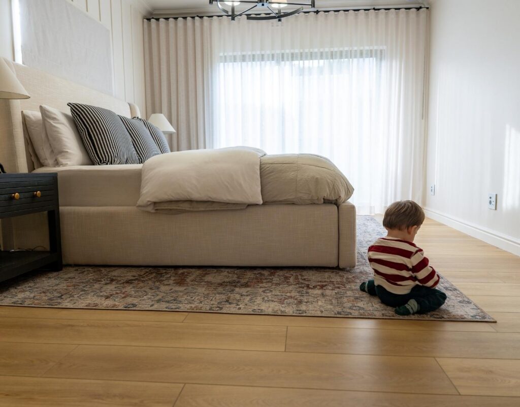 A young toddler in a striped shirt sitting on a patterned rug laid over light wood vinyl flooring in a spacious, neutral-toned master bedroom next to a large bed with a white headboard.